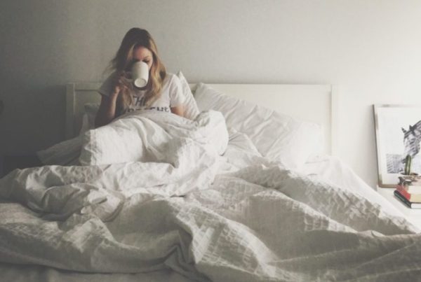 Woman under the covers in bed, hair in disarray, drinking a hot beverage in a mug