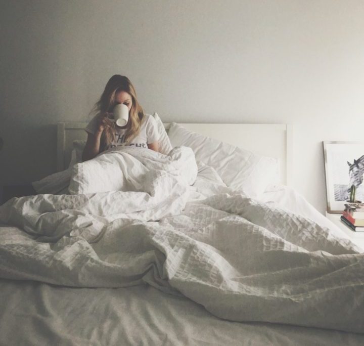 Woman under the covers in bed, hair in disarray, drinking a hot beverage in a mug