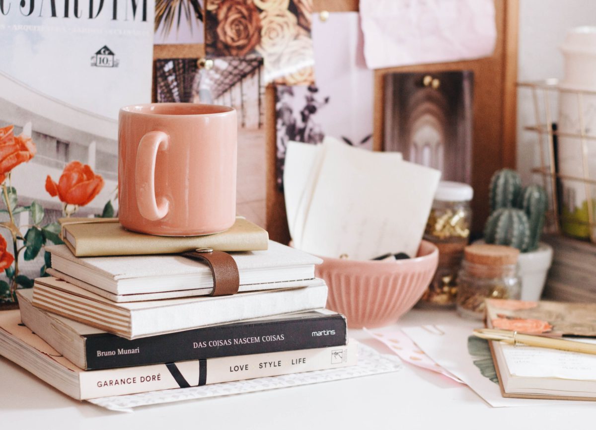 A work space featuring a pink aesthetic with a mug, several books, a vision board and cacti